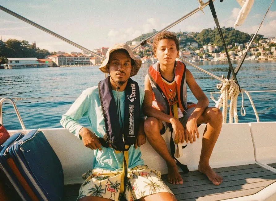 Skipper and a young sailor in lifejackets on deck, a harbour town behind them
